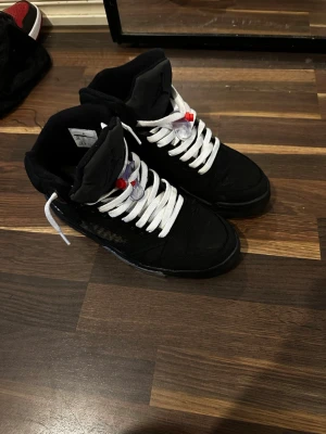 Nike Air Jordan 5 PSG Paris Saint-Germain - Säljer ett par svarta Nike Air Jordan 5 PSG Paris Saint-Germain sneakers med vita snören och detaljer i rött och grått. Skorna har meshpaneler med texten 'PARIS', broderad siffra 75 på hälen och reflekterande tunga. Ovandelen är i mocka och sulan har genomskinliga partier.