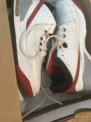 Nike Air Jordan 2 vit/röd sneaker - Nike Air Jordan 2 sneakers i vitt skinn med röda och svarta detaljer. Skon har perforerat mönster på ovandelen, snörning med röda öljetter och en synlig Air-enhet i sulan. Snygg retrostil med svart vadderad häl och grå inslag på mellansulan.