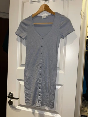 Ljusblå kortklänning från Vila - Kortklänning i ljusblått från Vila med små diskreta hålmönster, v-ringning och knappar hela vägen framtill. Klänningen har korta ärmar och är gjord i mjuk bomull som känns skön mot huden. Enkel och stilren modell som är lätt att matcha.