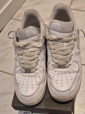 Nike Air Force 1 vita sneakers - Klassiska Nike Air Force 1 sneakers i helvitt skinn med perforerad tå, rund tå och snörning. Swoosh-logga på sidan och broderad Nike Air-detalj på hälen. Tjock, platt sula och vadderad krage för extra komfort.