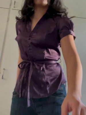 djuplila satintopp med knytning i midjan - En mycket elegant och feminin blus i en rik lila/plommonfärgad nyans. Toppen har en lyxig lyster tack vare satinmaterialet och en smickrande passform. Använt bara en gång. 36/S.