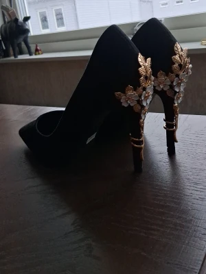 Svarta pumps med gulddetaljer och blommor - Säljer ett par svarta pumps från Eloshman i storlek 40. Skorna har spetsig tå och klack med unika guldfärgade metallblad och vita blommor som dekoration. Ovandelen är i mockaimitation och insidan är svart. Perfekta för dig som vill sticka ut! Dessa skor är enbart testade och säljs pga jag fick fel storlek när jag beställde. Orgpris 499kr.