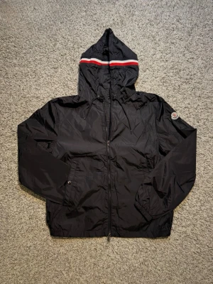 Moncler Windbreaker - Svart vindjacka från Moncler med huva och dragsko. Jackan har dragkedja framtill, två fickor med tryckknappar och en snygg Moncler-logga på ärmen (NFC koden fungerar). Huvan har en röd och vit rand som detalj. Perfekt för blåsiga dagar.