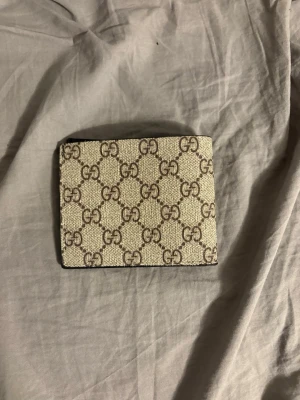 Gucci plånbok med GG-mönster - Säljer en snygg beige och brun plånbok från Gucci med det klassiska GG-mönstret på utsidan. Insidan är svart och har flera kortfack i skinn. Perfekt för dig som vill ha något stilrent och lyxigt i fickan.