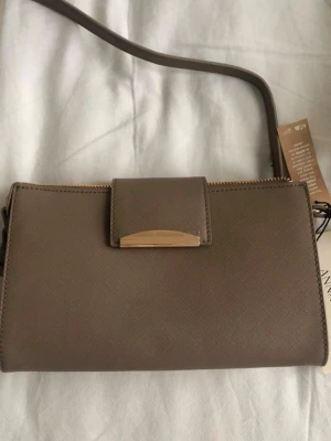 Taupe axelväska från Anna Field - Snygg taupefärgad axelväska från Anna Field i återvunnen polyester. Väskan har justerbar axelrem, guldfärgad detalj på locket och dragkedja upptill. Stilren och enkel design som passar till många outfits.