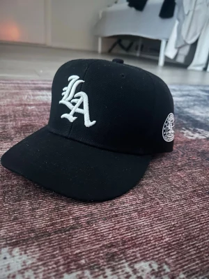Svart LA-keps med broderad logga - Säljer en svart keps med vit broderad LA-logga framtill och en rund Los Angeles California-patch på sidan. Klassisk rak skärm och stilren look som passar till streetwear. Kepsen är i ett tåligt tygmaterial och har en snygg kontrast mellan svart och vitt.