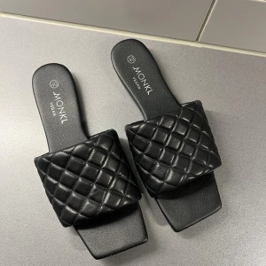 Svarta quiltade sandaler från Monki - Säljer ett par svarta sandaler från Monki med bred, quiltad rem i veganskt material. De har fyrkantig tå och platt sula, vilket ger en trendig look. Perfekta för sommaren och riktigt snygga till både jeans och klänning.