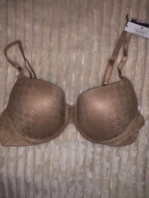 Bronsfärgad push-up bh Victoria's Secret - Säljer en snygg bronsfärgad push-up bh från Victoria's Secret, modell The Icon. Bh:n har tunna axelband och diskret logomönster över hela kuporna. Materialet är glansigt och känns lyxigt mot huden. Perfekt för dig som vill ha extra lyft och stilren look.