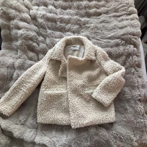 Beige teddyjacka från Stradivarius - Säljer en supermjuk och fluffig beige teddyjacka från Stradivarius. Jackan har klassisk krage, två fickor framtill och stängs med dolda knappar. Perfekt för dig som vill ha en cozy look med trendig vibe.
