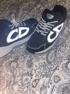 Svarta Dior B30 sneakers med reflekterande detaljer - Säljer ett par svarta Dior B30 sneakers med vita och reflekterande CD-detaljer på sidorna. Skorna har vit sula, meshpaneler och vita snören. Märket syns tydligt på plösen och hälen. Riktigt snygga och trendiga sneakers för dig som vill sticka ut.