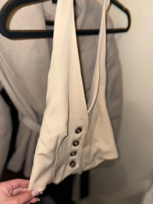 Beige croppad väst från Vero Moda - Säljer en beige croppad väst från Vero Moda med djup v-ringning och fyra snygga knappar framtill. Västen har breda axelband och elastisk detalj i ryggen för skön passform. Perfekt att styla över en skjorta eller t-shirt.