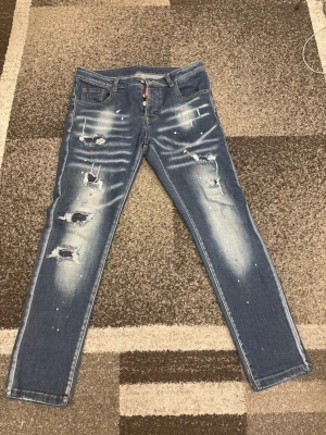 Dsquared2 blå distressed jeans - Säljer ett par blå jeans från Dsquared2 med coola slitningar, färgstänk och en stor röd logopatch bak. Jeansen har rak passform, klassisk femficksdesign och är tillverkade i slitstarkt denim. Perfekta för dig som gillar streetwear och edgy stil.