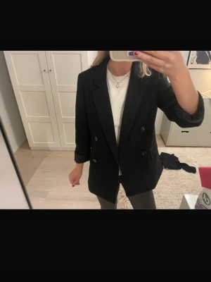 Svart kavaj - Säljer en svart kavaj med dubbelknäppning framtill. Klassisk krage och långa ärmar, perfekt att slänga över en t-shirt för en clean look. Tidlös och enkel att styla till olika outfits.