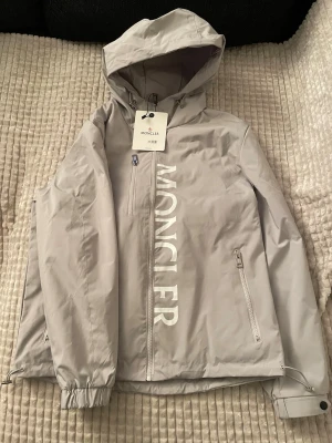Ljusgrå vindjacka från Moncler - Snygg ljusgrå vindjacka från Moncler med stor logga framtill, huva och dragkedjor på fickorna. Jackan har justerbar nederkant och är tillverkad i ett lätt, vattenavvisande syntetmaterial. Perfekt för dig som vill ha en clean och stilren look.