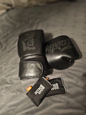 Svarta Fairtex boxningshandskar - Säljer ett par svarta boxningshandskar från Fairtex i skinn med kardborreknäppning. Handskarna har en stilren design med Fairtex-logga i svart och passar perfekt för träning eller sparring. Medföljer två SmellWell-doftpåsar för fräschör 14oz