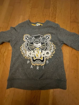 Grå Kenzo sweatshirt med tigertryck - Säljer en grå sweatshirt från Kenzo i storlek S med ikoniskt tigertryck och Kenzo Paris-logga framtill. Tröjan har rund halsringning och ribbade muddar. Perfekt statement piece för dig som gillar streetstyle och vill sticka ut.