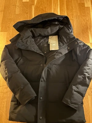 Svart dunjacka från Canada Goose M - Säljer en svart dunjacka från Canada Goose med avtagbar huva och pälsdetalj. Jackan har dragkedja, knappar och klassisk patch på ärmen. Perfekt för kalla dagar och riktigt snygg streetstil.