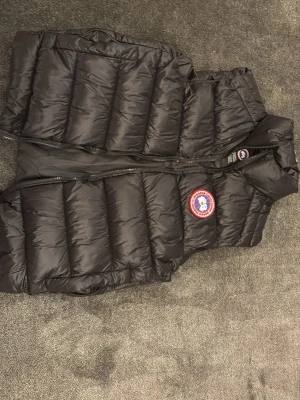 Svart dunväst från Canada Goose M - Säljer en svart, klassisk dunväst från Canada Goose med hög krage och dragkedja framtill. Västen har det ikoniska runda Canada Goose-märket på bröstet och breda, quiltade paneler som ger en riktigt schysst look. Perfekt för lager-på-lager under kyliga dagar.helt äkta påsen tillkommer också 