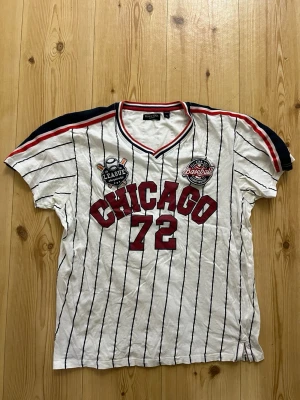 Randig t-shirt Chicago 72 Brave Soul - strl.XL - Säljer en vit t-shirt från Brave Soul med tunna svarta ränder och röda detaljer. På bröstet står det 'CHICAGO 72' i rött och det finns coola baseboll-inspirerade patches. Kragen har röd, vit och marinblå kant. Perfekt för en sportig vibe.