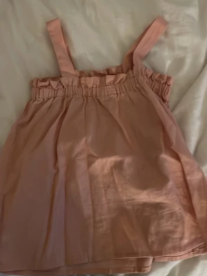 Ljusrosa linnetopp med volangkant - Supersöt ljusrosa linnetopp med breda axelband och volangkant upptill. Toppen har en avslappnad passform och är gjord i ett mjukt, tunt material som känns luftigt. Perfekt för varma dagar och enkel att styla med jeans eller kjol.