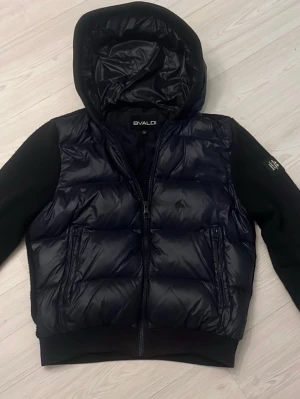 Blå/Svart pufferjacka från Bvaldi med huva - Säljer en svart/blå pufferjacka från Bvaldi i storlek M. Jackan har en stor huva, quiltad front och ärmar i mjukare material. Två dragkedjeförsedda fickor framtill och en snygg logga på ärmen. 