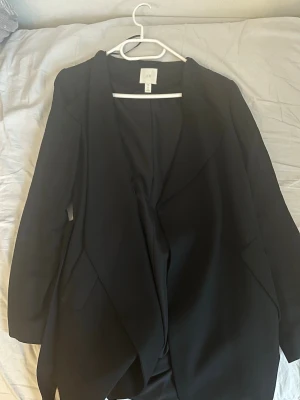 Svart kappa med bälte från H&M XS - Säljer en svart kappa från H&M i storlek XS. Jackan har ett snyggt bälte i midjan och klassisk krage. Långärmad modell med enkel, stilren look och fickor framtill. Perfekt för dig som gillar minimalistisk stil.