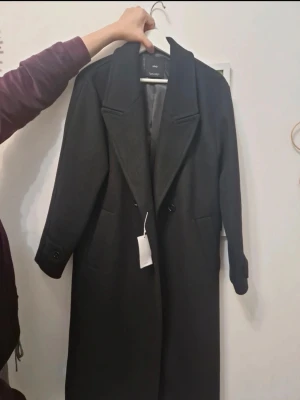 Svart kappa från Mango XS - Stilren svart kappa från Mango i storlek XS. Klassisk rak modell med bred krage, knappar framtill och fickor på sidorna. Perfekt för dig som vill ha en tidlös och elegant look. Materialet är mjukt och känns lyxigt. Helt ny med tags.