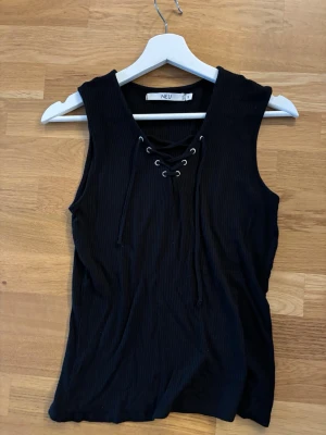 Svart ribbad linnetopp med snörning - Säljer en svart ribbad linnetopp från Lager 157i storlek S. Toppen har snygg snörning framtill och är ärmlös, vilket ger en cool och trendig look. Perfekt att matcha med jeans eller kjol för en avslappnad stil.