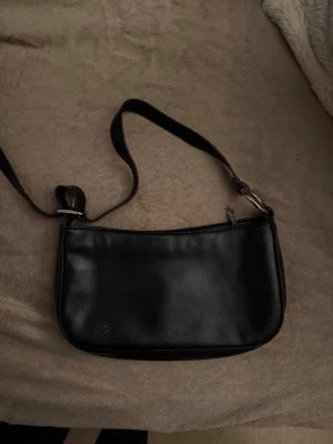 Svart axelväska - Snygg svart axelväska i skinnimitation från pull & bear med minimalistisk design och silverfärgade metalldetaljer. Väskan har justerbart axelband och stängs med dragkedja upptill. Perfekt storlek för det viktigaste och passar till många olika outfits.