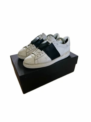 Valentino Garavani Open sneakers vit/svart - Säljer ett par Valentino Garavani Open sneakers i vitt skinn med bred svart läderdetalj över mitten. Klassisk rund tå, vita snören och platt sula. Märkeslogga på plösen och coola nitar bak på hälen. Perfekta för dig som gillar stilrena men ändå statement skor.