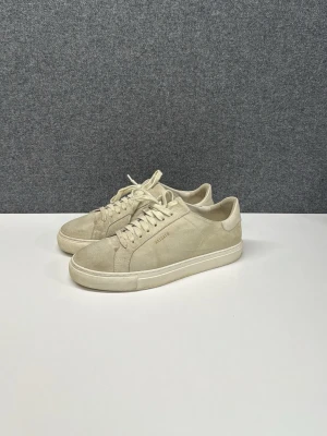Beige sneakers från Axel Arigato - Säljer ett par stilrena beige sneakers från Axel Arigato i mocka med klassisk låg profil och rund tå. Skorna har vita snören och diskret logga i guld på sidan. Perfekta till jeans eller chinos för en clean look. Skriv angående frågor!