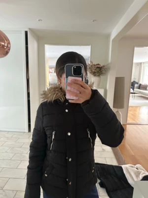 Svart pufferjacka med fuskpäls - Säljer en svart pufferjacka med huva och fuskpälsdetalj. Jackan har dragkedjor på fickorna och knappar framtill. Perfekt för kalla dagar och riktigt skön att bära. Klassisk modell som funkar till det mesta.