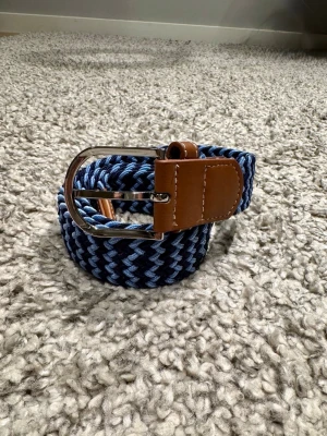 Blått Flätat Bälte - Ett stilrent flätat bälte med färgerna ljus- och mörkblå. Gjord av elastiskt material som anpassas efter behov. Spänner är gjort av läder och metall. Längden är 105 cm