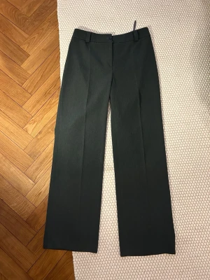 Mörkgrå kostymbyxor, storlek 40 - Säljer ett par mörkgröna kostymbyxor i klassisk rak modell med hög midja och bälteshällor. Byxorna har pressveck framtill och är sydda i ett mjukt, vävt material som ger en snygg och stilren look. Perfekta till en snygg topp eller skjorta. Jag är 167