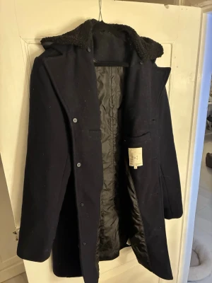 Mörkblå ullkappa från Tailored Originals - Säljer en mörkblå kappa från Tailored Originals i storlek XL. Skulle säga att den passar en M/L. Kappan har en avtagbar krage i teddy, knäppning framtill och praktiska fickor. Materialet är ullblandning och insidan är fodrad för extra värme. Perfekt för kyliga dagar och riktigt snygg till höst- och vinterlooken.