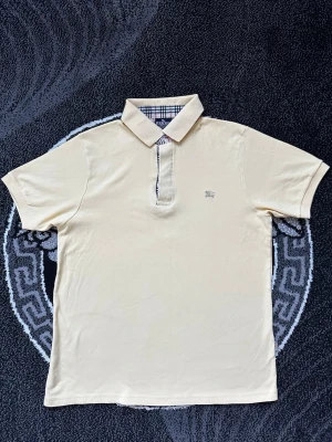 Beige Burberry piké med rutiga detaljer - Säljer en klassisk beige pikétröja från Burberry London med ikoniskt rutigt mönster på insidan av kragen och knappslån. Pikén har korta ärmar, broderad Burberry-logga på bröstet och är gjord i mjuk bomull. Perfekt för en clean och stilren look.