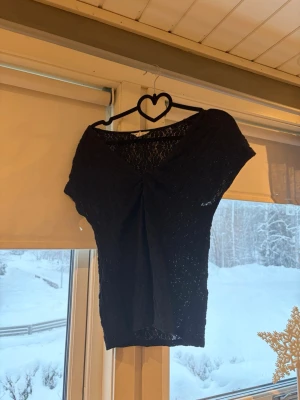 Svart spetsblus från Dobber - Säljer en svart blus från Dobber i storlek M. Toppen har v-ringning, korta ärmar och är gjord i ett genombrutet spetsmaterial med blommönster. Snygg twistdetalj framtill som ger en unik look.