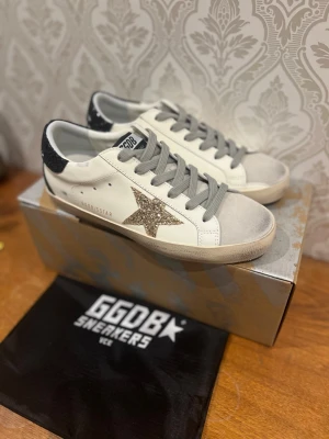 Golden goose skor - Snygga GGDB sneakers i vitt skinn med grå snörning och beige mockadetalj på tån. Sidan pryds av en glittrig stjärna i guld och hälen är svart med glitter. Klassisk låg modell med platt sula och cool vintagekänsla.