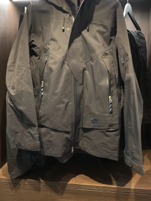 Svart skidjacka från State of Elevate - Säljer en svart vindjacka från State of Elevate med huva och flera dragkedjeförsedda fickor. Jackan har justerbara muddar med kardborre och snygga detaljer på insidan. Perfekt för dig som vill ha en stilren och funktionell jacka.