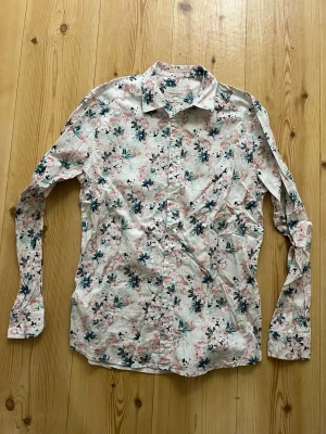 Blommig slim fit skjorta Tailored Originals - Säljer en blommig skjorta från Tailored Originals i slim fit. Skjortan är vit med rosa och gröna blommor, har långa ärmar och knappar framtill. Tillverkad i mjuk bomull med lite stretch för extra komfort. Perfekt för dig som vill sticka ut lite extra.