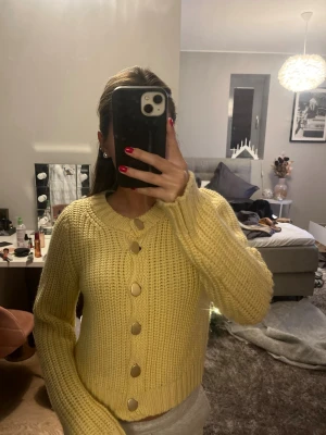 Gul stickad kofta med stora knappar - Superfin gul stickad kofta med stora, runda guldknappar framtill. Koftan har en chunky känsla, ribbade muddar och lång ärm. Perfekt för att liva upp din outfit med en poppig färg och mysig vibe.