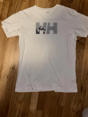 Vit t-shirt från Helly Hansen - Vit t-shirt från Helly Hansen med stor HH-logga och bergmotiv på bröstet. Klassisk passform och rund halsringning. Tillverkad i mjuk bomull som känns skön mot huden. Perfekt för en avslappnad stil. Finns täcken på användning på loggan, enbart där.