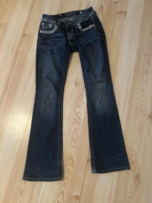 Miss Me bootcut jeans med broderi - Säljer ett par mörkblå Miss Me jeans i bootcut-modell, low waist. Jeansen har snygga broderade detaljer och nitar på fickorna samt kontrastsömmar. Materialet är klassisk jeans med stretch. Perfekta för dig som gillar unika detaljer och cool stil. Lite slitet på ena knäet. Se sista bilden 