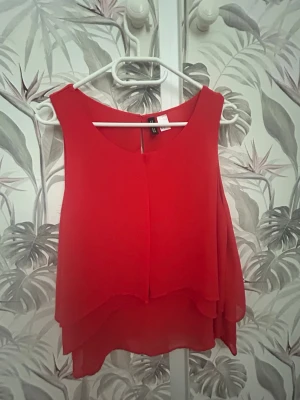 Röd omlott linnetopp  - Säljer en röd ärmlös linnetopp från H&M med dubbla lager i luftig polyester. Toppen har rund halsringning och ett snyggt omlottfall framtill som ger en flowy look. Perfekt för dig som vill ha en enkel men ändå statement topp.