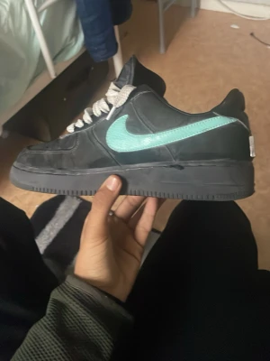 Nike Air Force 1 x Tiffany & Co svart - Säljer ett par Nike Air Force 1 x Tiffany & Co i svart skinn med turkos Swoosh och silverdetalj på hälen. Klassisk låg modell med vita snören och platt sula. Riktigt clean och unik sneaker för dig som vill sticka ut.