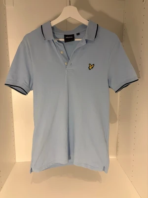 Ljusblå piké från Lyle & Scott - Säljer en ljusblå pikétröja från Lyle & Scott med klassisk krage och knappar framtill. Pikén har svarta detaljer på krage och ärmslut samt den ikoniska gula fågel-loggan broderad på bröstet. Tillverkad i bomull för en skön och luftig känsla.