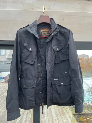 Woolrich Jacka - Säljer en marinblå fieldjacket från Woolrich med flera praktiska fickor och snygga tryckknappar. Jackan har hög krage, dragkedja och detaljer i metall. Materialet är slitstark polyester och jackan har en klassisk, tidlös look.