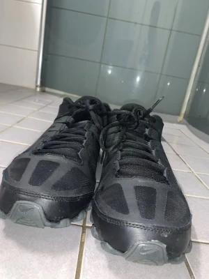 Nike Reax svarta sneakers - Säljer ett par svarta Nike Reax sneakers med cool design och robust sula. Skorna har meshpaneler för bra andningsförmåga, syntetdetaljer och tydliga Nike-loggor på sidan och tån. Perfekta för dig som gillar sportig stil och vill ha något bekvämt till vardags.