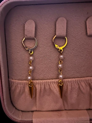 Guldiga örhängen med pärlor och spikes - Säljer ett par snygga örhängen i guld-look med små vita pärlor på kedja och spetsiga spikes längst ner. Perfekta för dig som vill ha en edgy men ändå elegant vibe. Passar till både vardag och när du vill sticka ut lite extra.