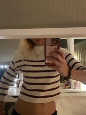 Randig croppad tröja - Säljer en croppad stickad tröja med vita och mörkröda ränder. Tröjan har trekvartsärm och ribbad kant vid hals, ärmslut och nederkant. Perfekt för dig som gillar en avslappnad men trendig stil.